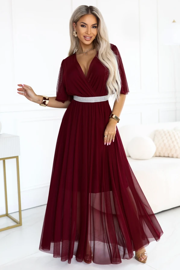 Modele bordo krāsas Numoco Basic maxi kleitā ar krustotu izgriezumu, caurspīdīgām piedurknēm, sudrabainu rotaslietām līdzīgu jostu un platinātu tīkliņa apakšdaļu virs īsākas oderes.