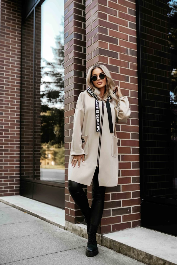 Modelka prezentuje beżowy płaszczyk damski z kapturem i ozdobnymi taśmami „LIFE IS BEAUTIFUL” marki Numoco Basic.