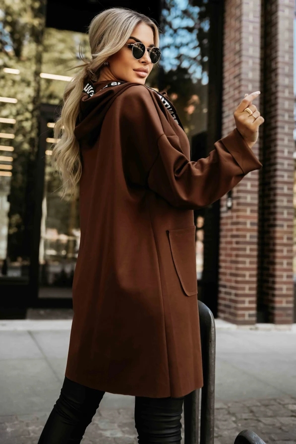 Modelka prezentuje stylowy płaszczyk w kolorze czekoladowym z napisami na troczkach, podkreślając jego luźny i nowoczesny krój – Numoco Basic.