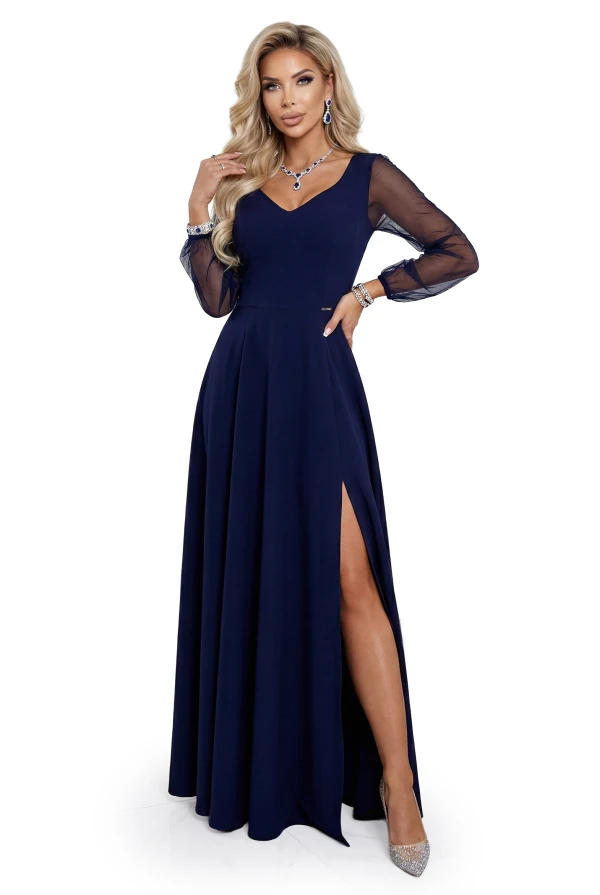 Modelka w eleganckiej, granatowej sukni maxi z dekoltem V, transparentnymi długimi rękawami i rozcięciem na nodze, prezentująca wieczorową stylizację na wesele, galę lub przyjęcie w jasnym, minimalistycznym wnętrzu.