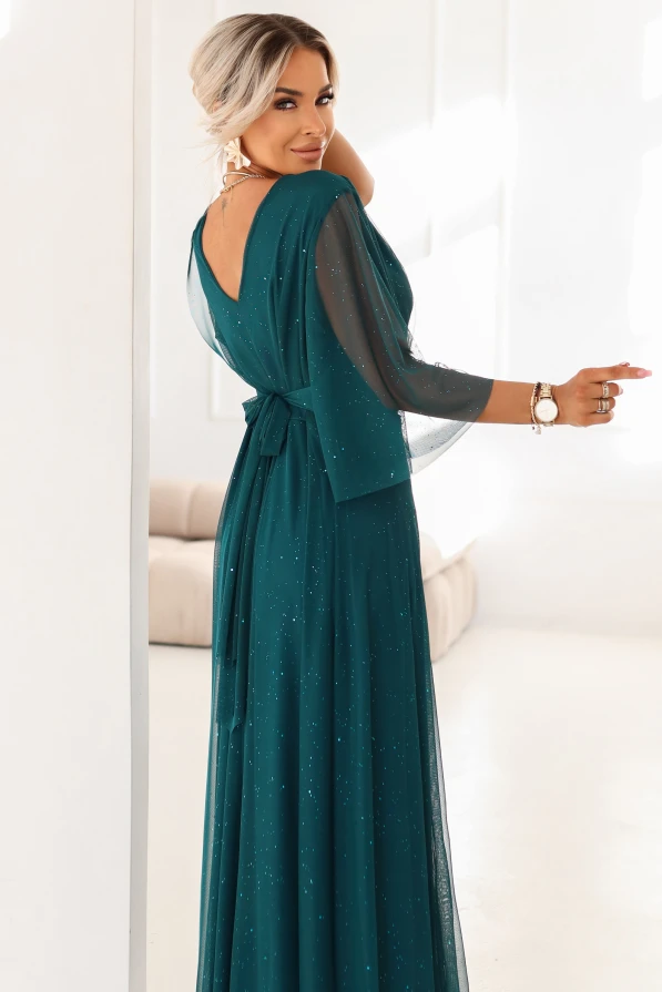 Wieczorowa długa sukienka plus size w butelkowej zieleni z lekkim tiulem i brokatem – modelka stoi w jasnym, pałacowym wnętrzu.